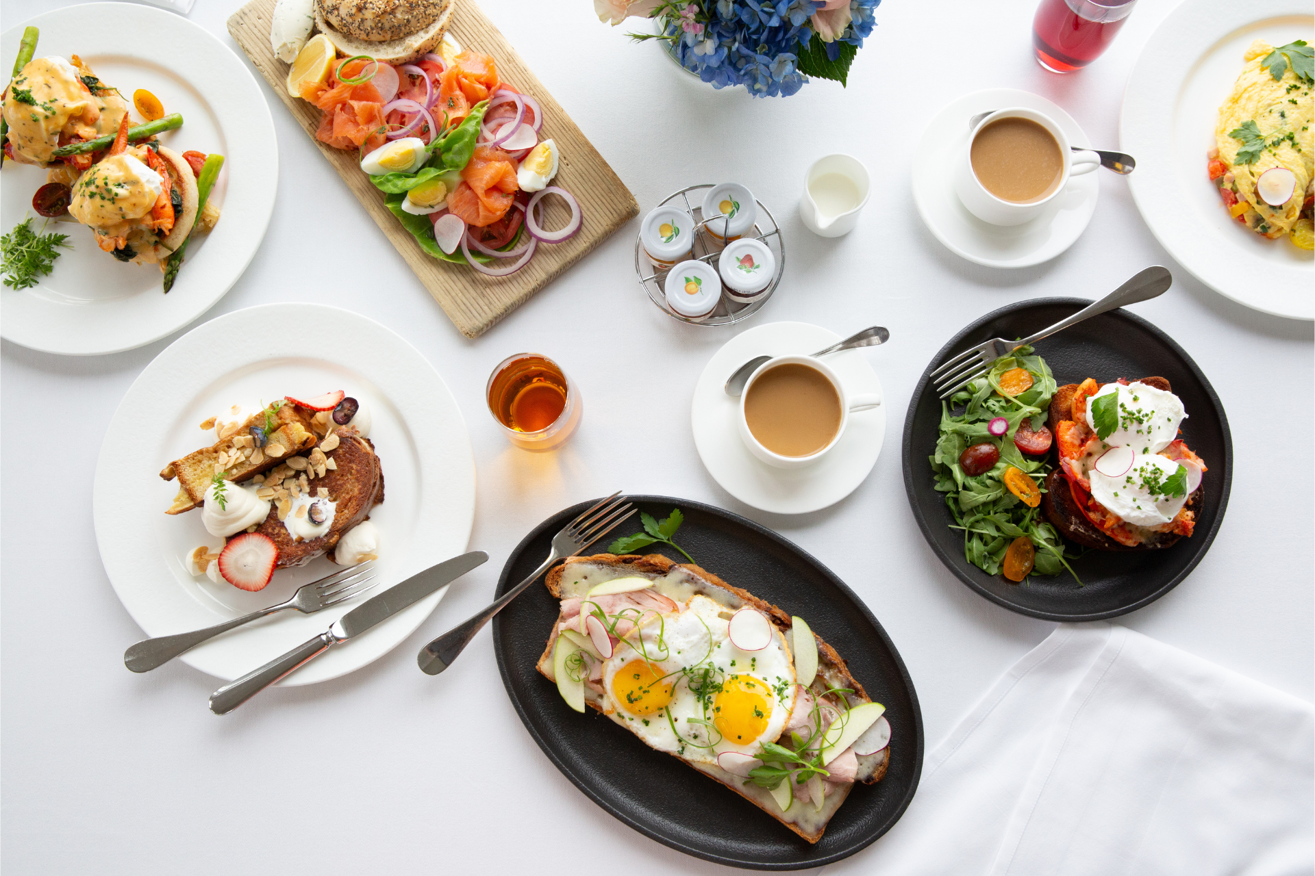 A variety of breakfast dishes including eggs, salmon, coffee, juices, toast, salad, and a floral centerpiece on a white table.