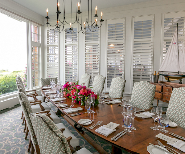A dining room with a long table set for a meal, featuring flowers, elegant chairs, a chandelier, and a decorative sailboat on a sideboard.