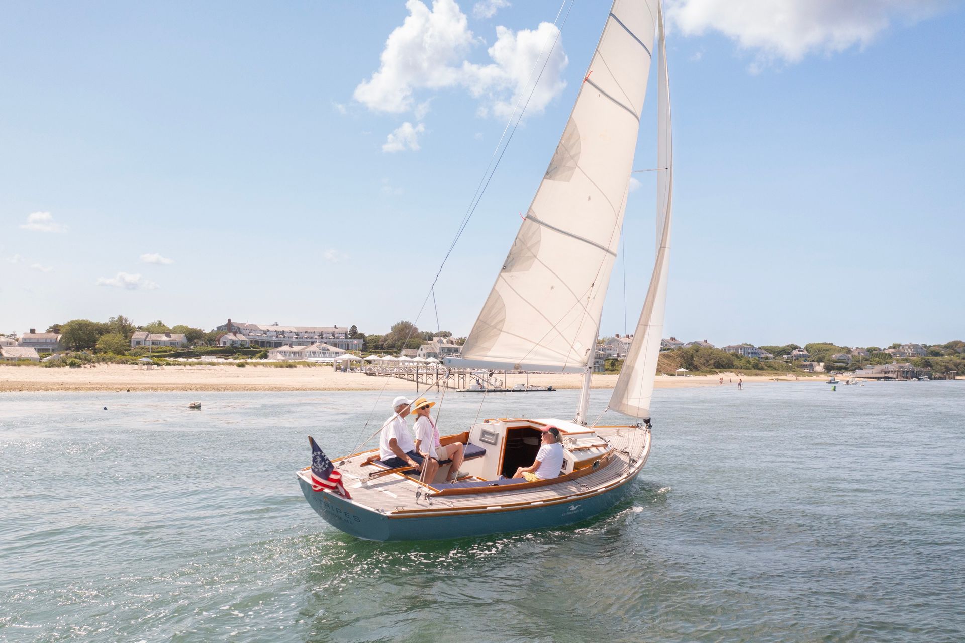 A sailboat with three people is on the water near a sandy beach with houses in the background under a clear blue sky.