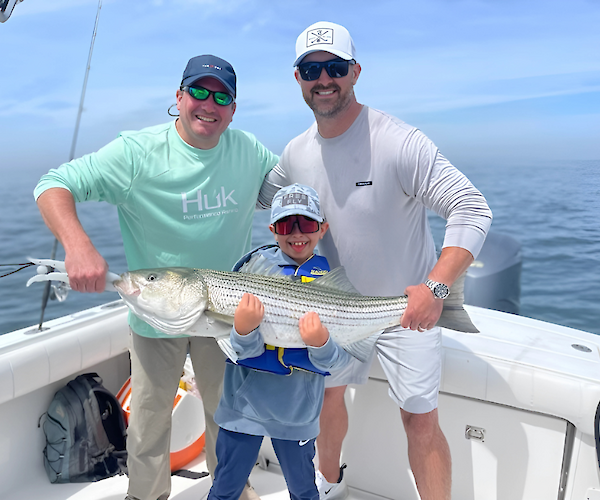 Three people on a boat holding a large striped fish, smiling and dressed in casual fishing gear.