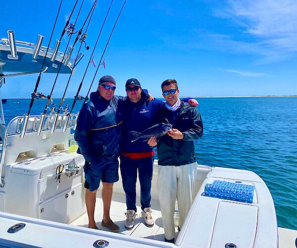 Three people on a boat with fishing rods; one holds a fish. Clear blue sky and water in the background.