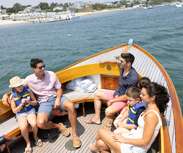 A family enjoys a sunny day on a boat, with blue waters and a shoreline in the background.