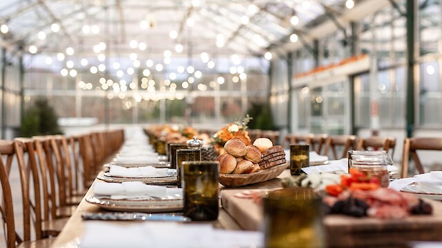 A long table set for a meal in a greenhouse, with bread, glasses, and plates, under soft string lights, creating an elegant atmosphere.