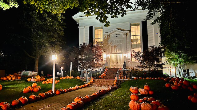 A well-lit building with a grand staircase, surrounded by numerous pumpkins arranged decoratively on the pathway and lawn.
