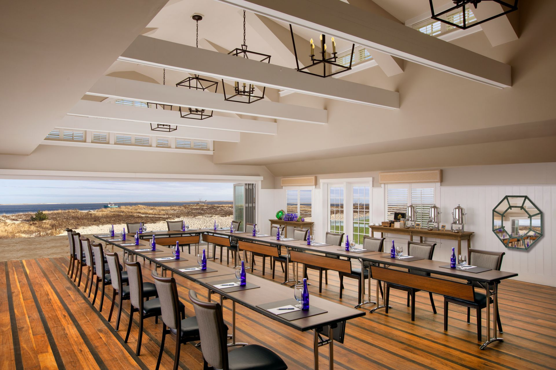 A conference room with arranged tables and chairs, blue bottles on each table, overlooking a coastal landscape through large windows.