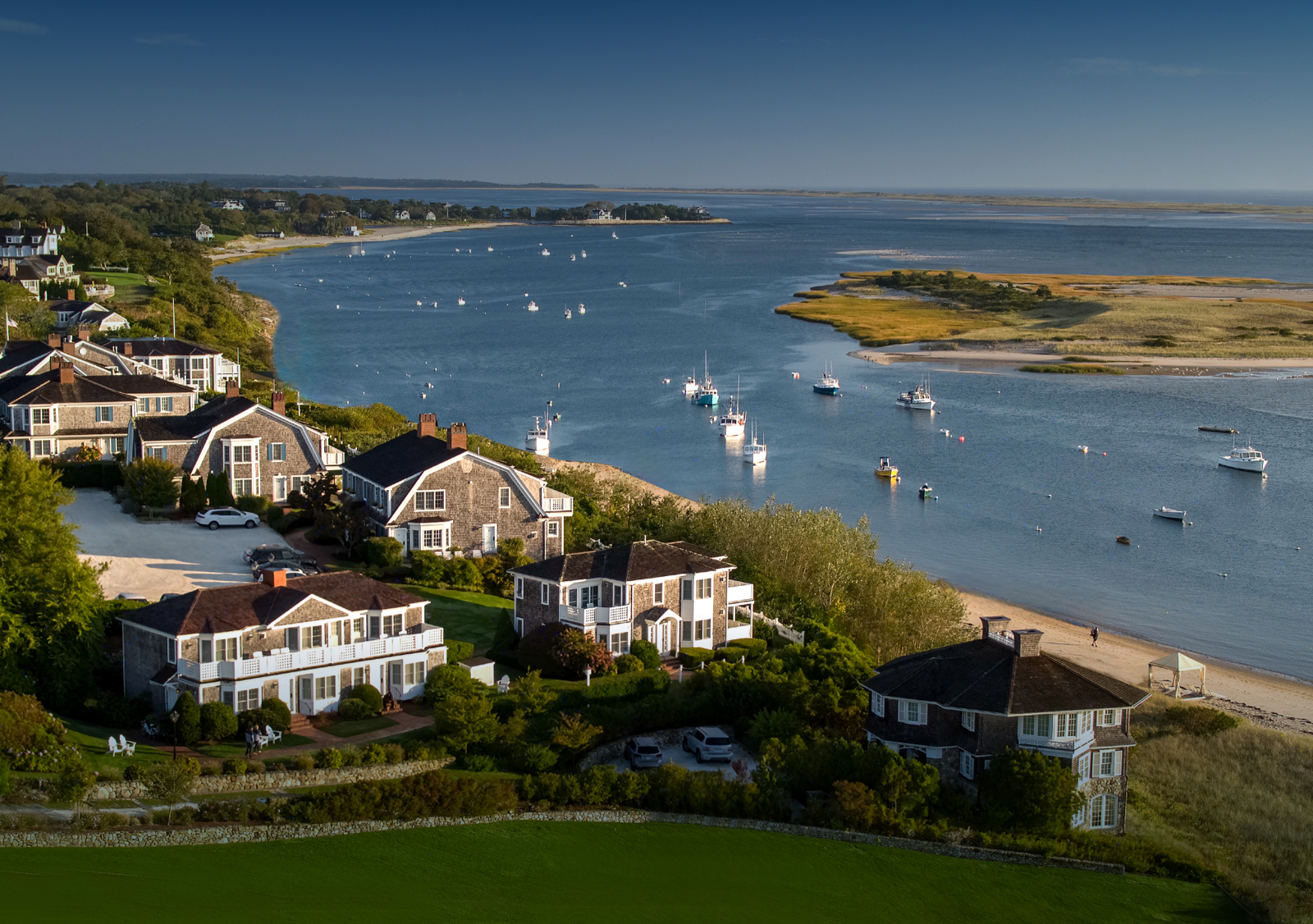 Coastal scene with waterfront homes, a sandy beach, and boats on a calm bay under a blue sky.