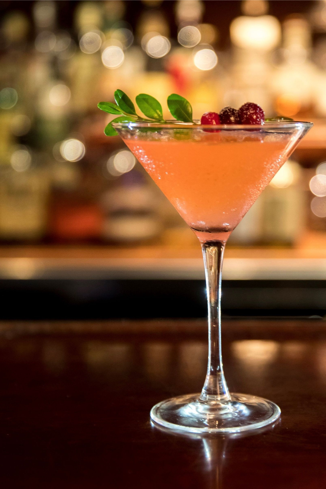 A cocktail in a martini glass garnished with berries and a sprig of herbs, set against a blurred bar background.