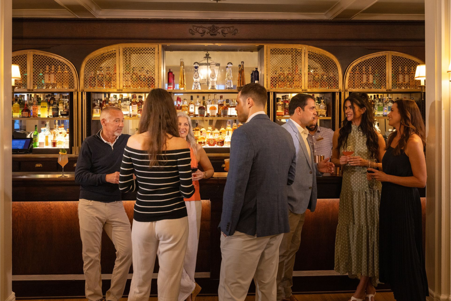 People socializing in a warmly lit bar, dressed in smart casual attire, with shelves of liquor bottles in the background.