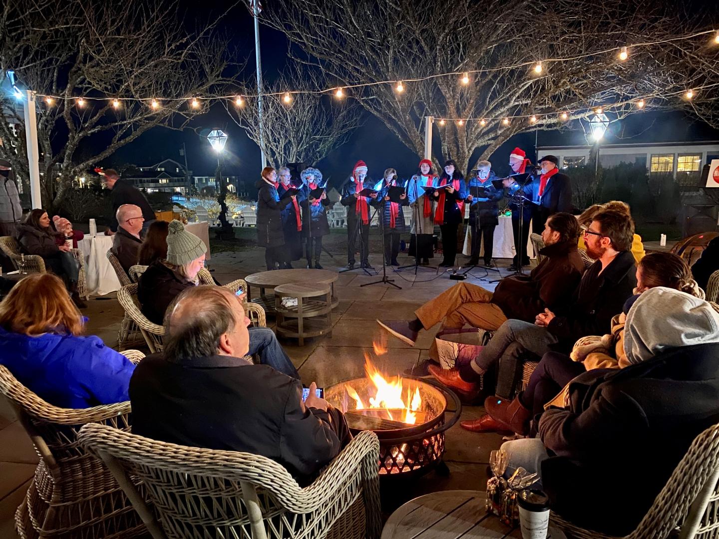 Carolers sing while guests gather around an outdoor firepit drinking hot cocoa